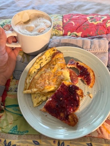 Photo of a plate with an omelette beside toast & jam. Beside the plate a hand holds a white coffee mug filled with a spiced latte with foam.